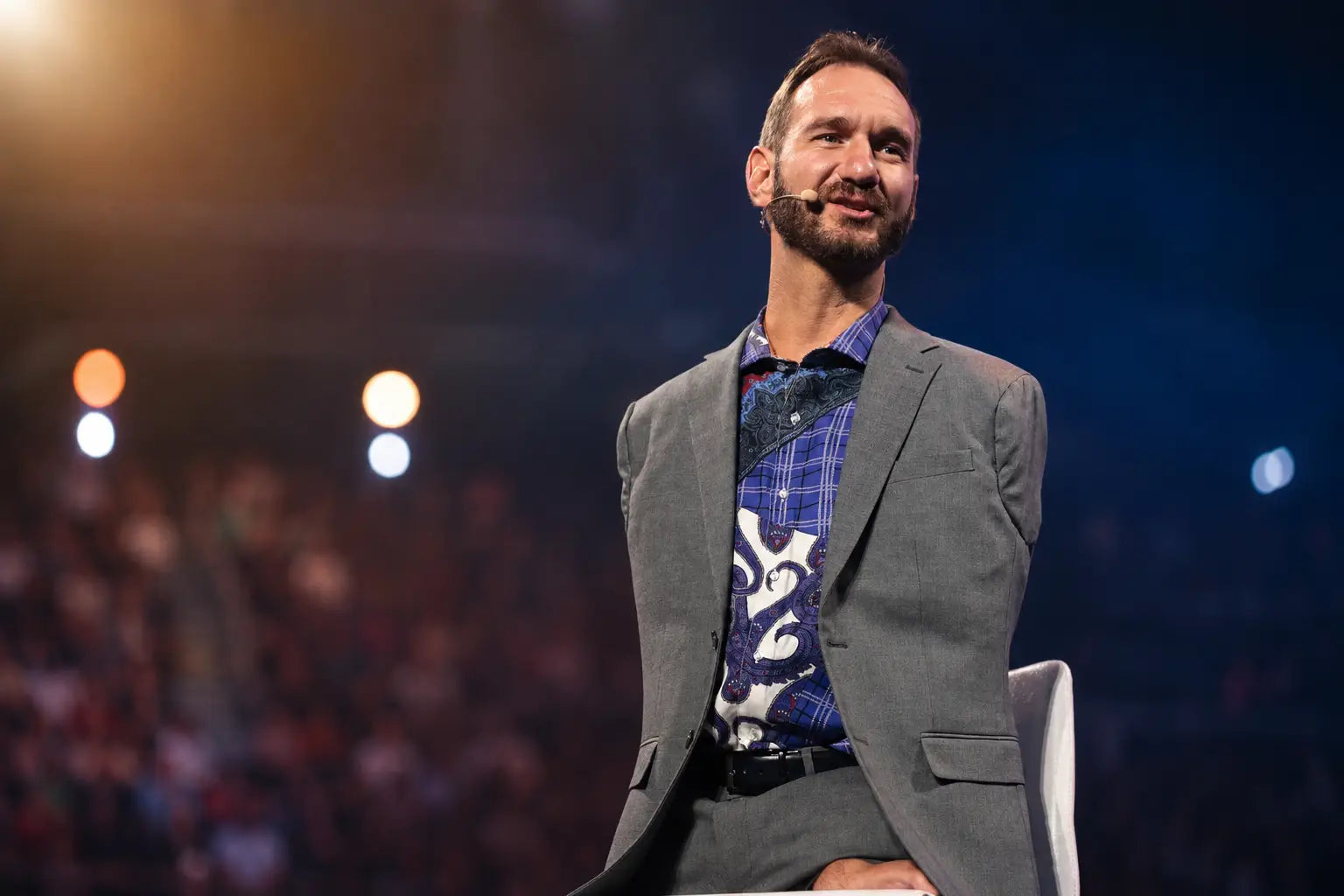 Nick Vujicic seated on a stage in a gray suit and patterned shirt, speaking into a headset microphone with a blurred audience and warm stage lights in the background.