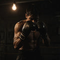 A lone boxer in a dimly lit gym stands under a single overhead light, gloved fists raised — the raw focused hunger of someone who trains when no one is watching.
