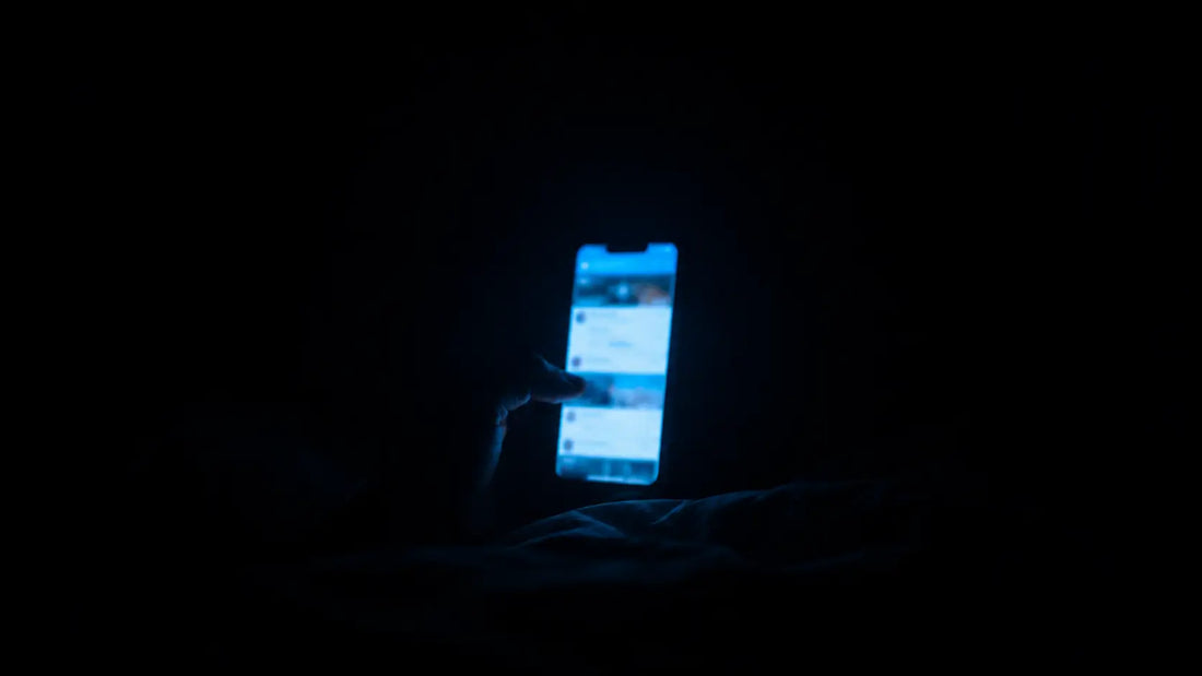 A phone screen casting blue light in a completely dark bedroom with a blurred social media feed visible, the glow the only illumination in the room, symbolising the doomscrolling loop
