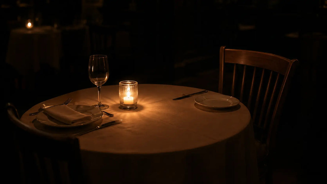 A small round restaurant table set for two in warm candlelight, one place setting with a wine glass and folded napkin as if someone arrived, the opposite chair empty with its place setting untouched