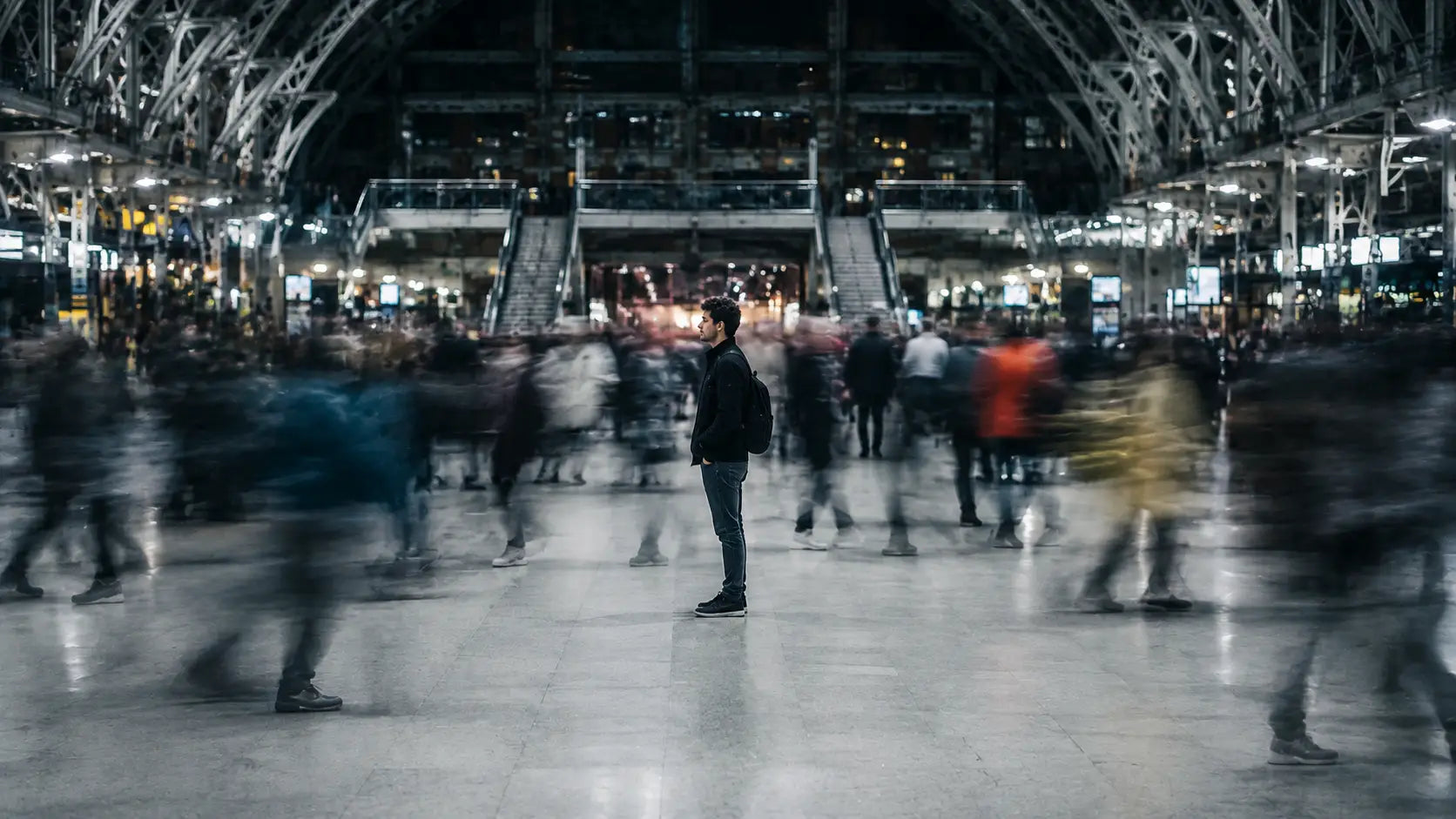 A person standing completely still in the centre of a busy train station concourse, every other person blurred into streaks of motion around them, the lone figure sharp and frozen amid the chaos